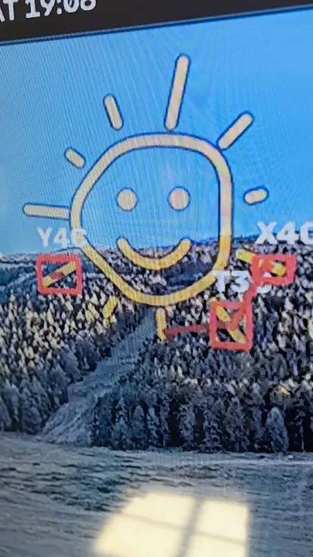 Photo of the stream on a screen: a hand-drawn smiling sun over the forested slope, with red detection boxes on the tips of its rays